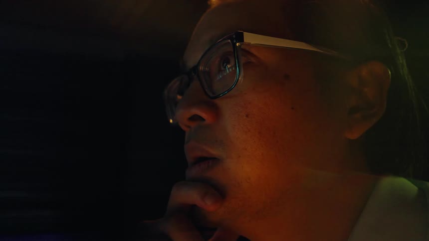 A close view of a man with glasses looking at a glowing computer screen in a dark room features heavy motion blur and light leak effects
