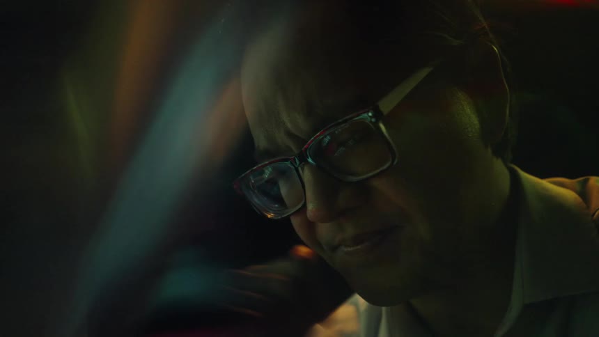 A man wearing glasses is shown in dramatic colorful lighting leaning forward with his hand near his mouth in a thoughtful pose as he removes his eyeglasses looking stressed