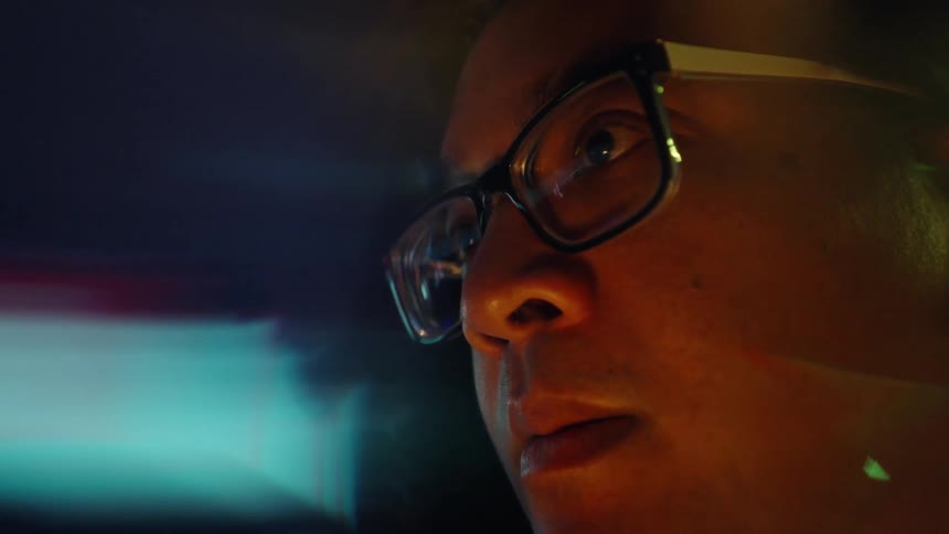 A man wearing glasses looks intently at a computer screen that reflects orange and blue financial bar charts onto his lenses while digital data visualizations blur in the dark background around his face