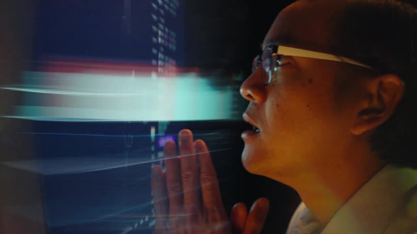 A man wearing glasses gazes intently at a glowing digital screen colorful light reflecting across his face as he clasps his hands thoughtfully