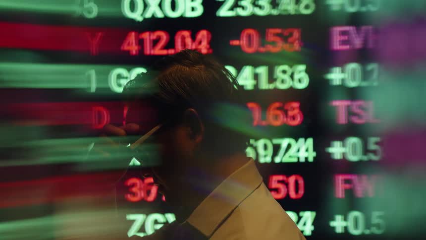 A side view of a man tired wearing glasses and a white shirt shows him next to a large screen filled with scrolling red and green stock market symbols and numbers in a dark room