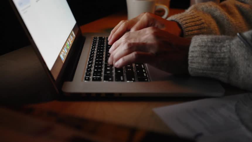 A person in a cozy sweater types on a laptop at a wooden desk with a coffee mug and papers nearby in warm ambient lighting