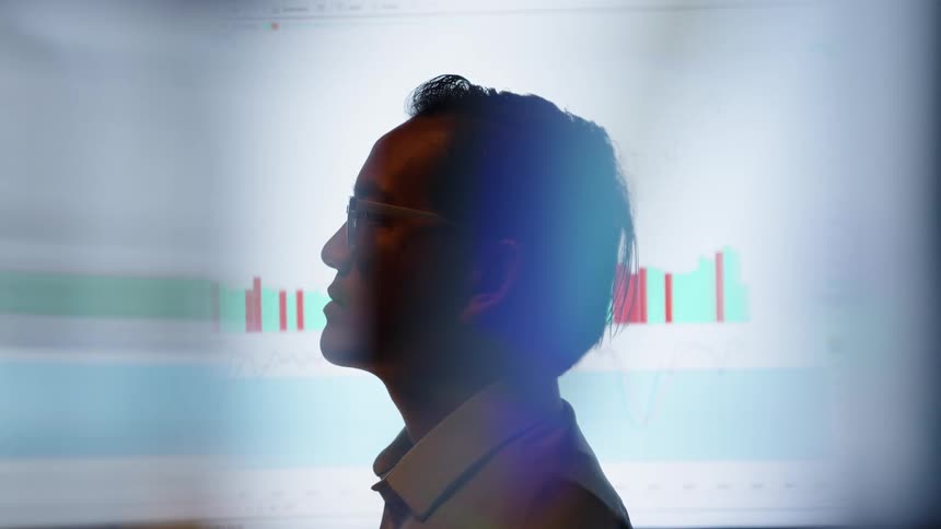 Man is briefly partially silhouetted against a monitor displaying charts