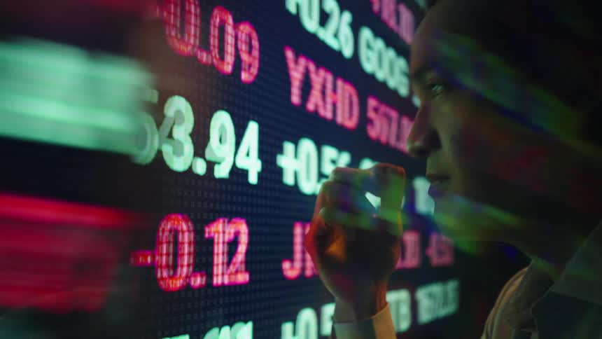 The video depicts a stressed man rubbing his eyes while standing in front of a glowing digital stock market ticker he eventually puts on his glasses to closely examine the fluctuating financial data and red numbers displayed on the screen