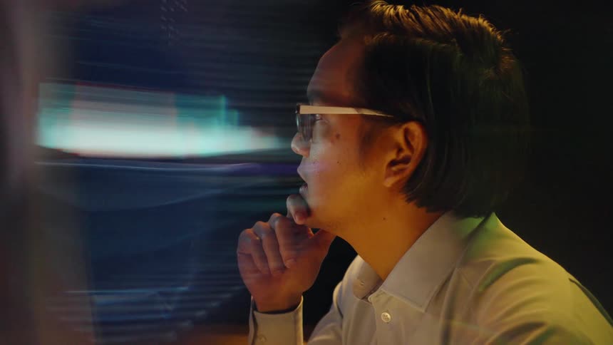 Man wearing glasses looks at an illuminated screen with hands clasped