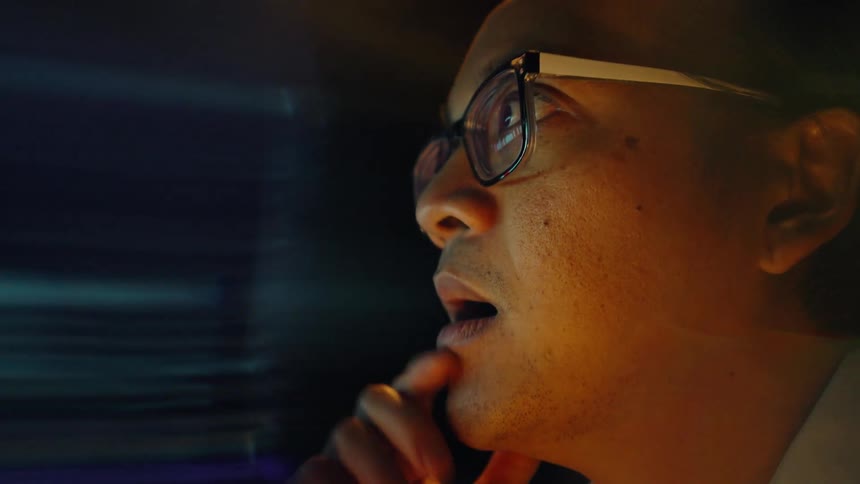 Adult man wearing glasses sitting and looking with a nervous facial expression at a financial graph on a screen indoors