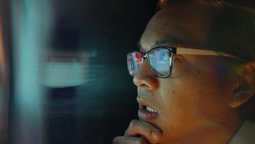 Man wearing glasses looks at illuminated screen with hand resting on chin