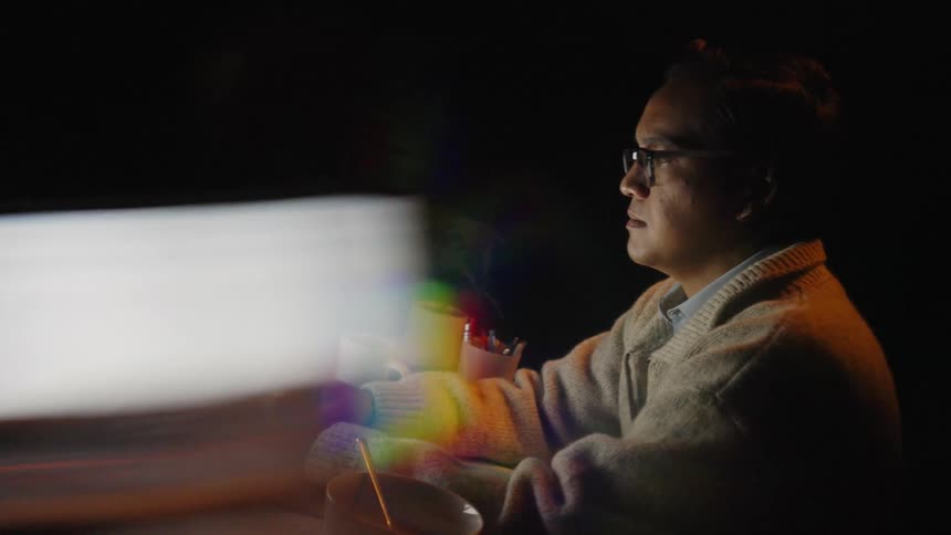 A man wearing glasses and a warm cardigan sits at a desk in a dark room focused on his laptop screenthe workspace is cluttered with a bowl mugs and papers all partially illuminated by the laptops glow and colorful lens flares