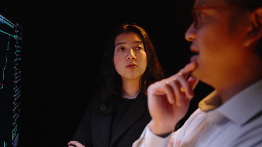 Man and woman standing in a room talking with computer monitors on the wall with graphic charts