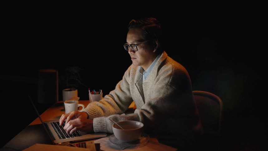 A man is sitting as he types on a laptop in the dark