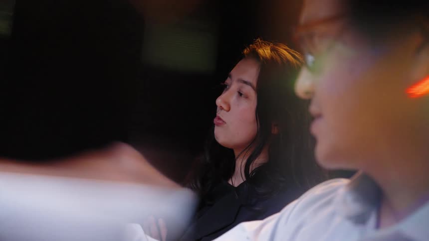 A woman and a man in professional attire stand in a dark room while she points at a glowing digital screen displaying colorful stock market data and candlestick charts to analyze financial trends
