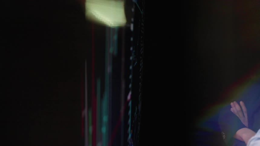 A woman with her arms crossed stands beside a colleague while looking at a large digital display the screen shows a financial candlestick chart with red and green bars which someone points to during the discussion