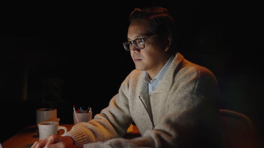 Man works on a laptop at night at a desk with a cup and documents