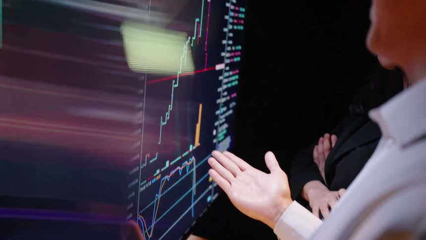 Two people in a dark room analyzing a large screen displaying realtime financial stock market charts and a man points at the fluctuating data trends while a woman stands beside him observing the technical indicators closely
