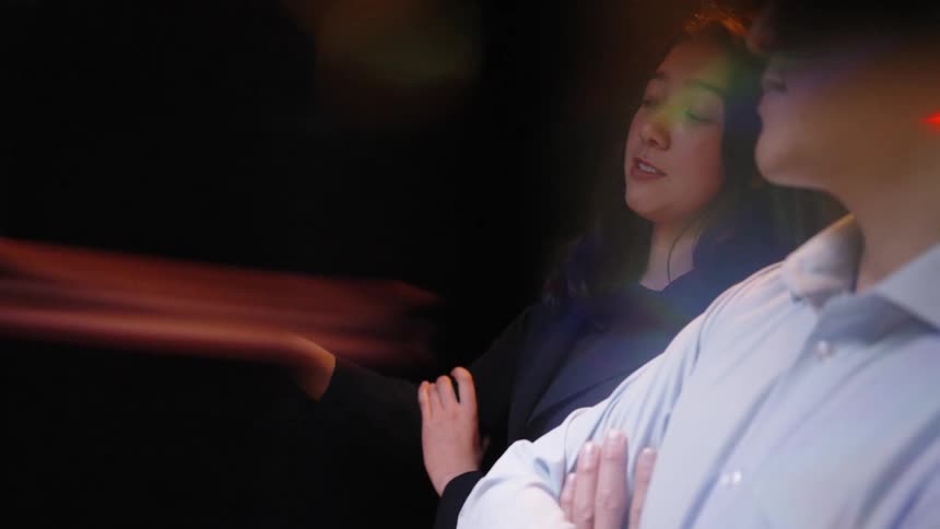 Woman speaks while a man with crossed arms listens both illuminated by colorful screen reflections in a dark setting