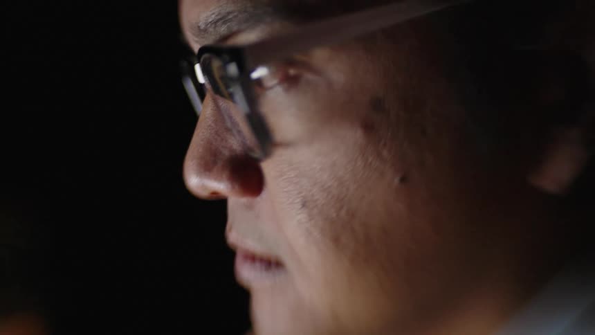 Man dons his eyeglasses then looks at the monitor reflected in their lenses at night