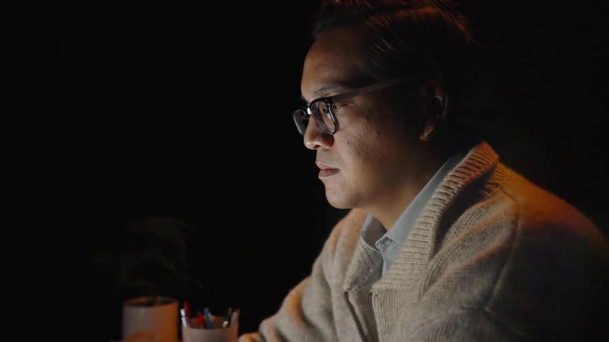 The video shows a man with glasses and a sweater working at a laptop in a dark room he appears focused as his face is illuminated by the screens glow while he looks at the monitor