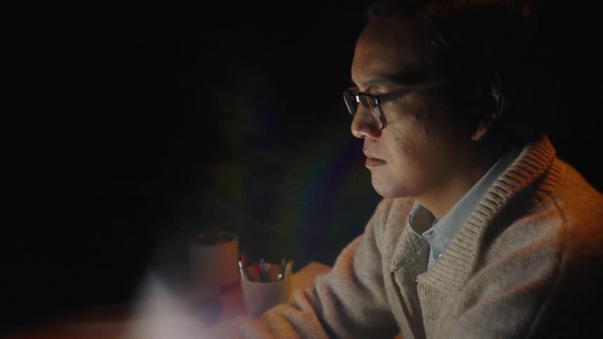 An asian man wearing a cardigan and glasses sits at a dark desk while typing on a laptop