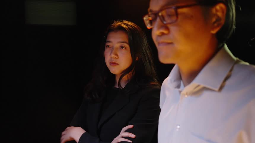 In a dark room a man and a woman in business clothes stand together while looking at a screen that provides a warm glow on their faces