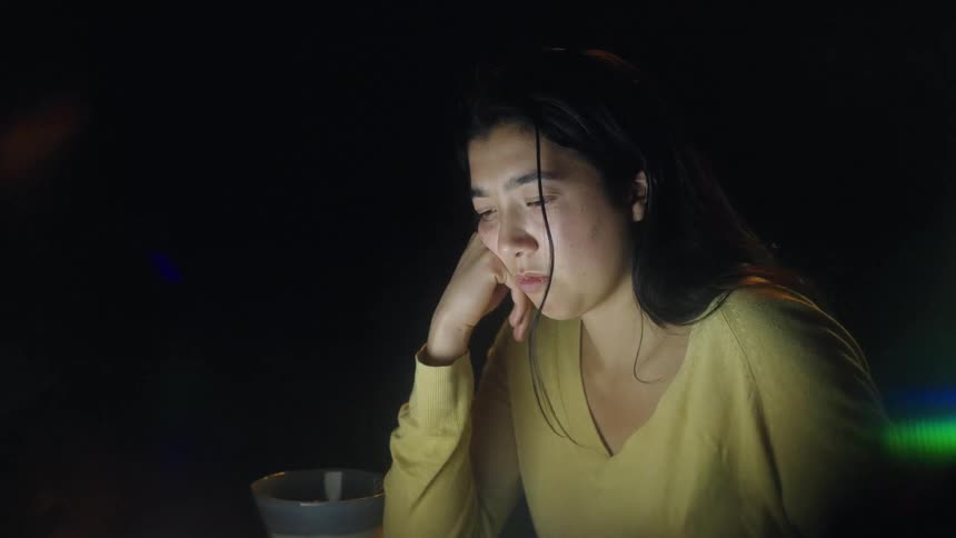 Woman resting her head on her hand looks at an illuminated screen in a dark environment