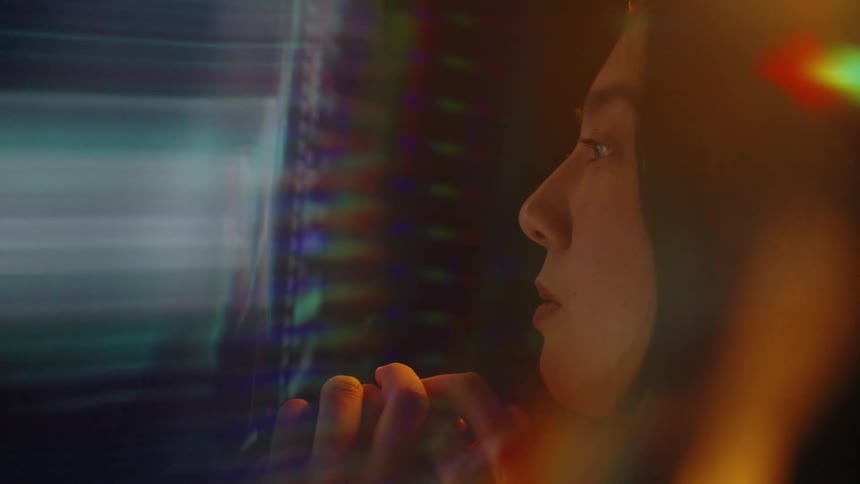 Young adult woman sitting with a contemplative facial expression surrounded by blurred colors and shapes indoors