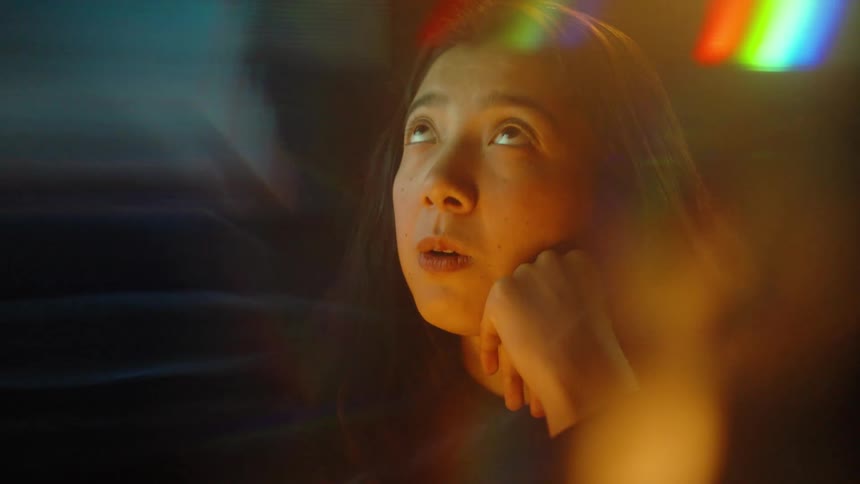 A woman sits in a dimly lit room with her chin resting on her hand appearing deep in thought or weary as light streaks across the frame she eventually looks upward with a curious or contemplative expression highlighted by colorful lens flares and a cin