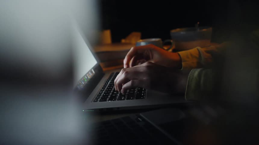 Hands typing on a laptop with cups in the background in a dark environment