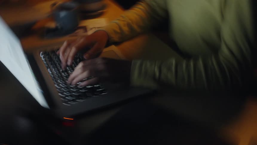 A close view shows the hands of a woman wearing a yellow sweater typing quickly on a laptop keyboard at a wooden desk in a dark room before the camera moves to her face as she looks down at the screen