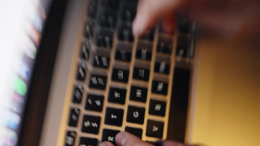 A closeup shot shows hands typing rapidly on a backlit laptop keyboard in a dimly lit environmentthe camera angle shifts slightly throughout the clip capturing the motion blur of the fingers and the glow of the screen reflecting off the
