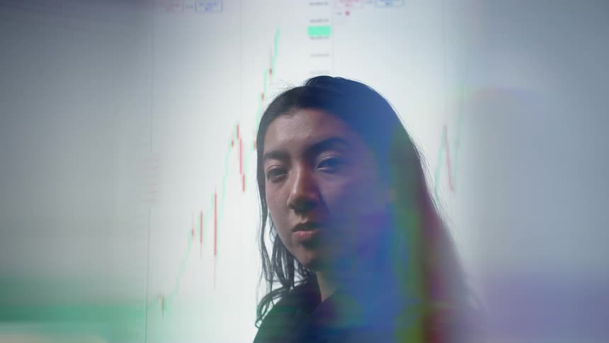 A woman with dark hair stands in profile as a white screen with a red and green stock market bar chart is visible through her silhouette with soft blue and purple light reflections in the foreground