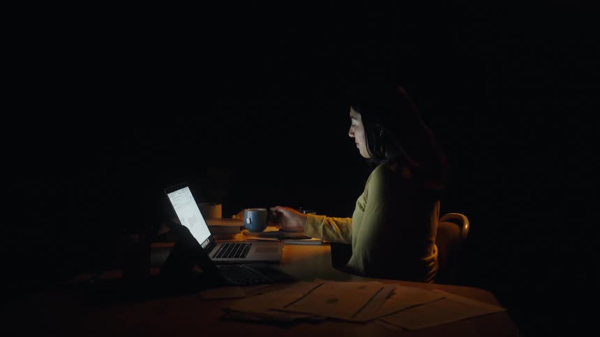 In a dark room a woman sits at a desk and drinks from a blue mug while working on a laptop and tablet surrounded by papers and a bowl