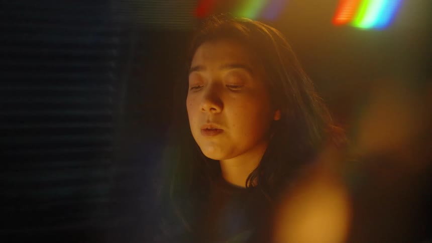 A close view of a woman in a dark room focuses on her as she looks at a screen that provides a warm glow on her face while bokeh and light leak effects blur the frame