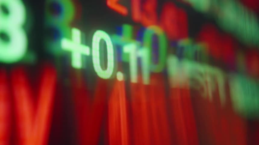 A close view of a glowing computer screen displays rapidly changing digital stock market symbols and numbers with a heavy motion blur and chromatic aberration effect