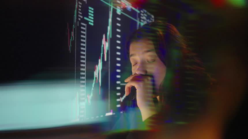 A woman with long dark hair sits in front of a computer screen while colorful financial charts and market data are projected across her face and the room