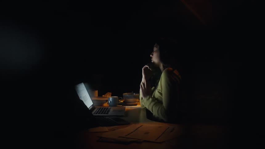 A woman wearing a yellow sweater sits at a desk in a dark room using a laptop then covers her face with her hands and rubs her neck while papers and a bowl sit nearby under a single light source