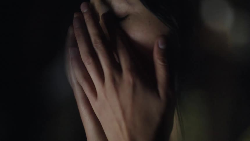 A woman wearing a yellow long sleeve shirt types on a laptop in a dark room then stops to rub her neck and face with her eyes closed as soft light reflects on her skin
