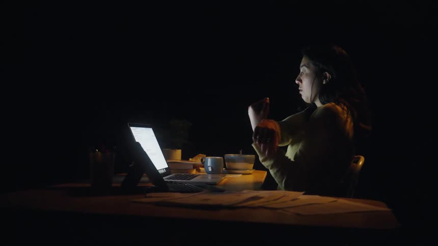 A woman wearing a yellow sweater sits at a desk and stretches her arms while working on a laptop in a dark room illuminated by the screen light