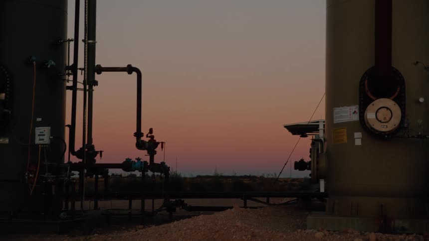 Industrial tanks and piping stand silhouetted against a pastel sunset sky in a flat open landscape