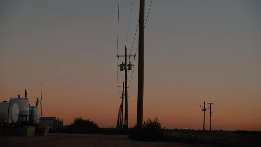 Power lines and utility poles stand against a sunset sky with industrial tanks in the foreground