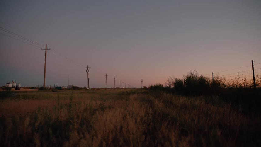 This video depicts a tranquil rural landscape at dusk featuring a line of utility poles stretching toward a fading horizon the golden hour light casts soft shadows across the tall grass and fenced fields creating a peaceful cinematic atmosphere