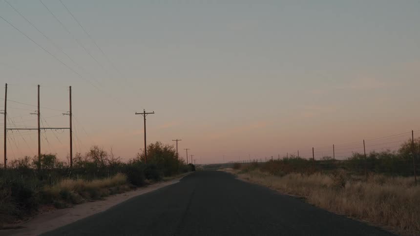 A narrow asphalt road stretches toward a distant horizon at twilight flanked by a series of wooden utility poles and power lines that cut across a soft fading orange sky over a quiet rural landscape
