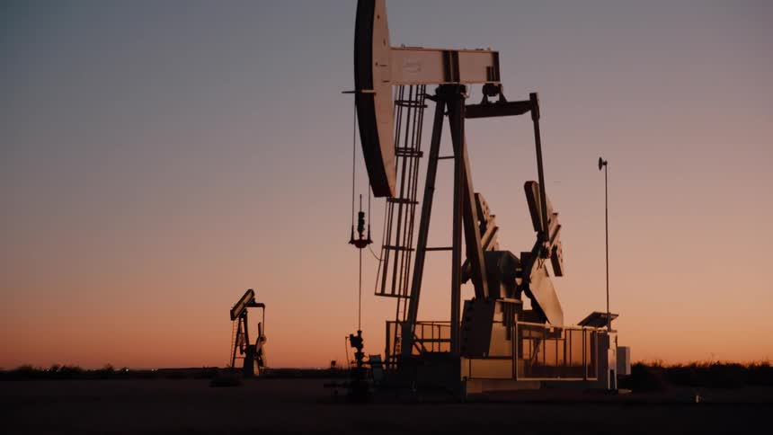 An industrial oil pumpjack rhythmically pivots up and down against the glowing backdrop of a desert sunset casting a sharp silhouette as the fading light paints the wide horizon in deep oranges and soft purples