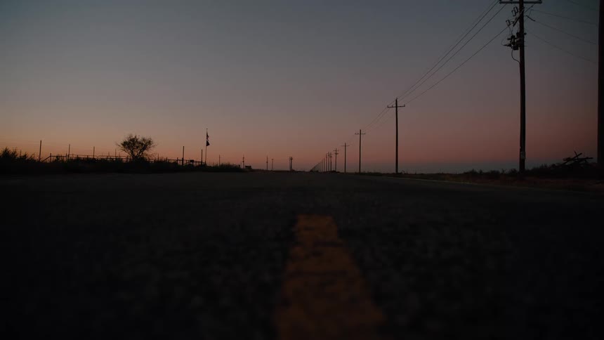 A low angle view shows a long asphalt road with a yellow center line stretching toward a distant horizon under a dark orange and blue twilight sky while wooden utility poles and a wire fence line the side of the road