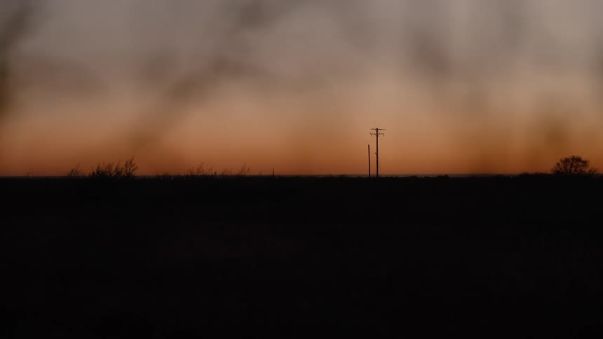 A large open area in the wilderness with electrical power lines and smoke rising into the atmosphere at sunset
