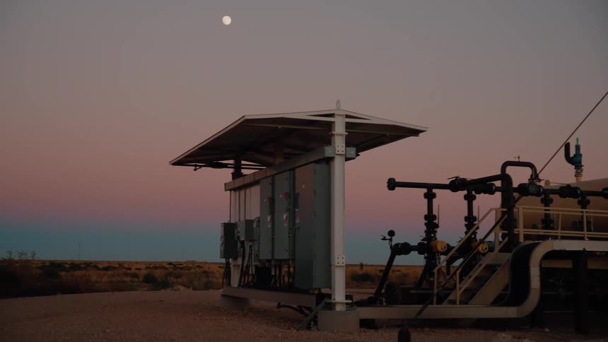 Industrial equipment sits under a wide pale sky with the moon visible