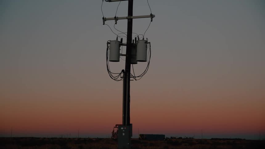 A utility pole with mounted transformers and cables stands against a gradient sunset sky over a flat landscape