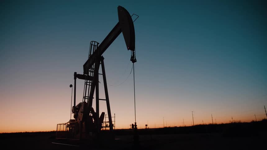 An oil pump jack operates in silhouette against a gradient sky during dusk or dawn