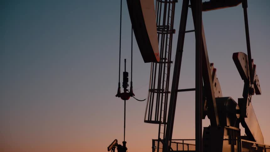 A metal pump jack operates in a dark field against a clear orange and blue twilight sky during a quiet sunset in an industrial area