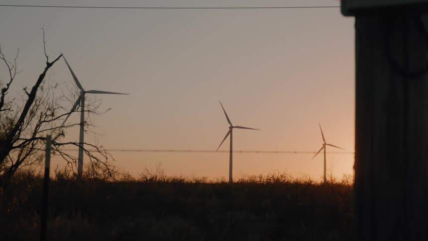 Three wind turbines rotate slowly in a field at sunset silhouetted against an orange and purple sky