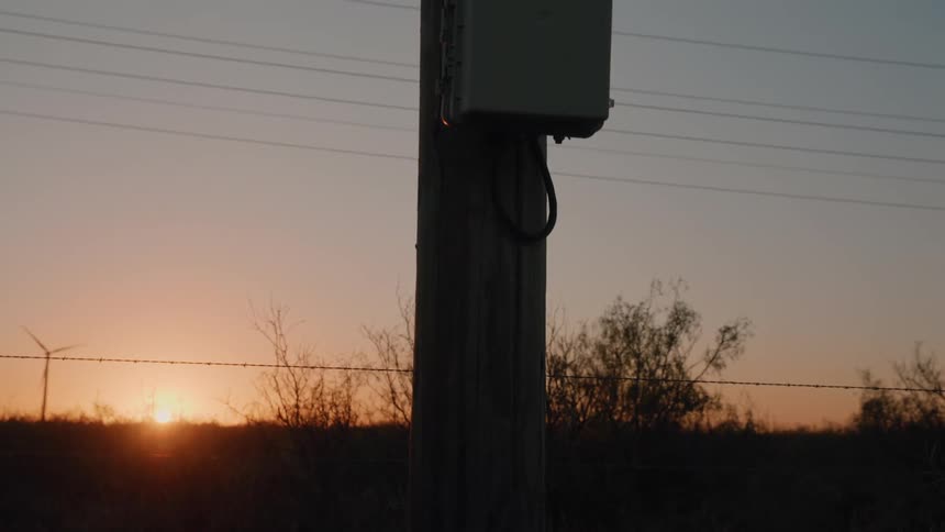 Up a wooden utility pole to reveal a grey control box as the sun sets on the horizon casting a warm orange glow over a rural landscape dotted with wind turbines and wire fencing